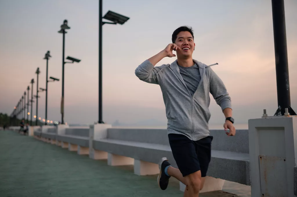 Man smiling and touching his ear while running in shorts and sweatshirt along a boardwalk. 